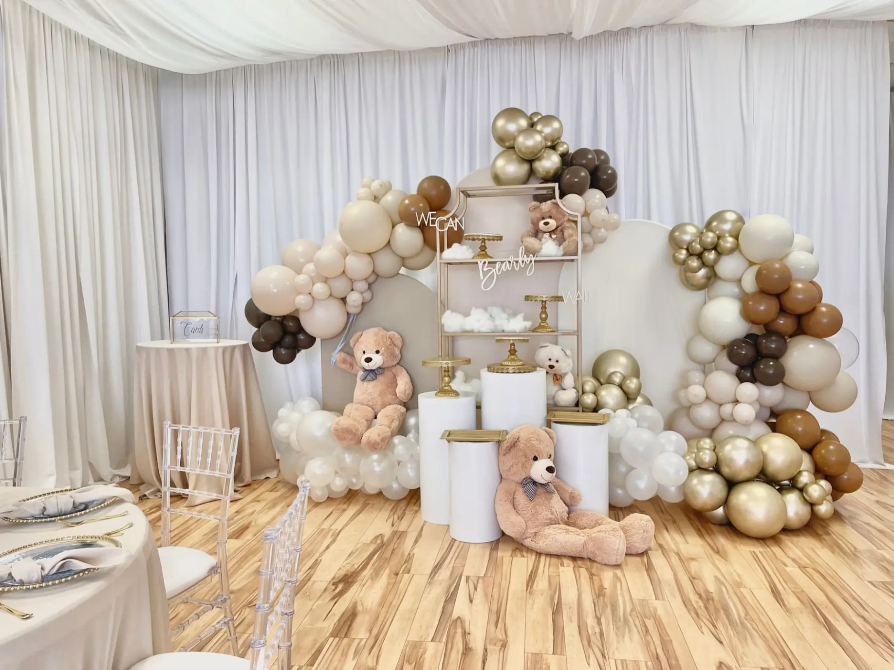 Neutral baby shower setup with white balloon arch, teddy bear décor, gold shelf and cylinder pedestals